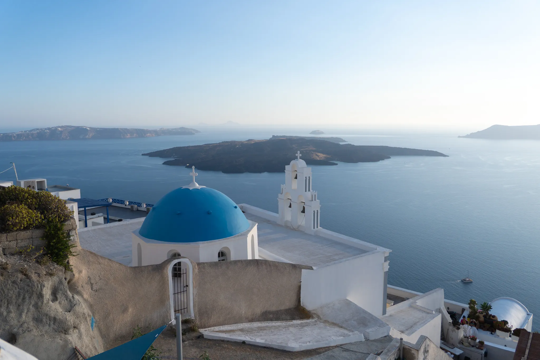 A blue-domed church and its white bell tower above the Santorini caldera at golden hour